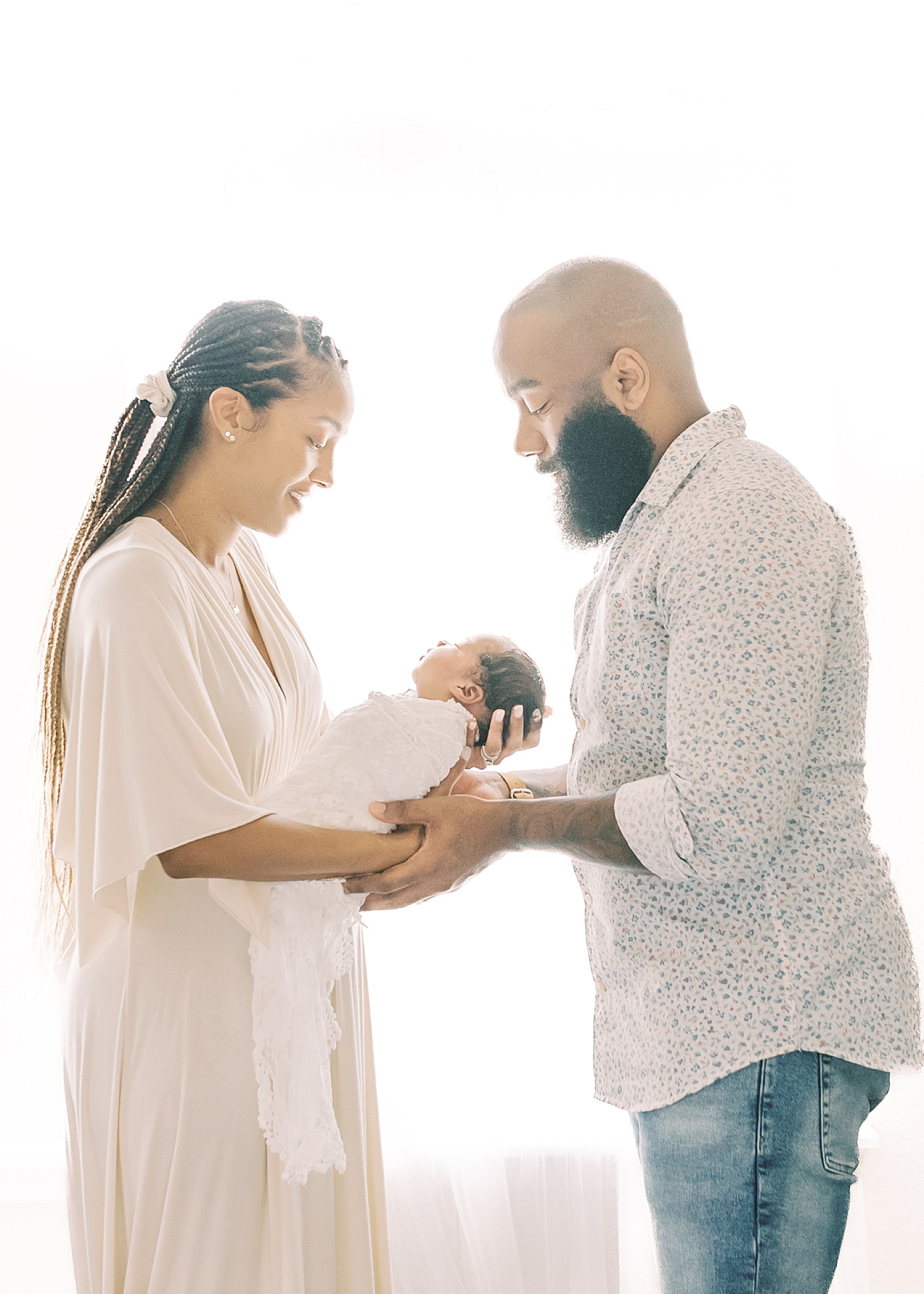 black man and black woman holding a newborn baby girl wrapped in a white lace wrap in front of an airy window