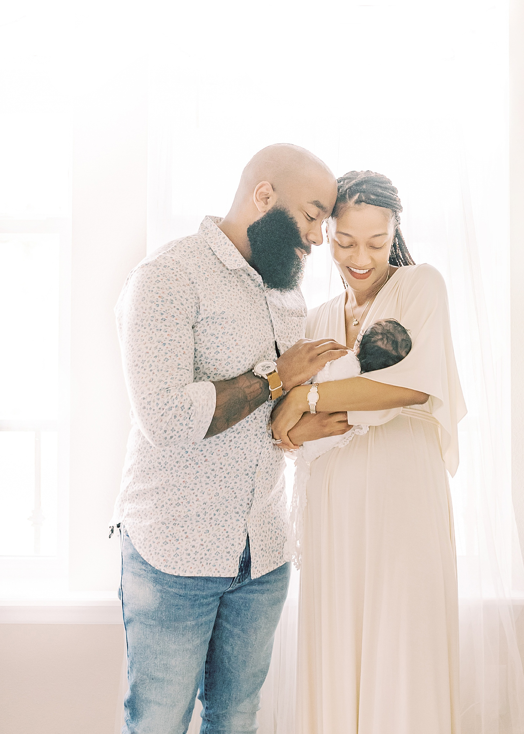black man and black woman holding a baby girl wrapped in a white lace blanket in front of an airy window with white sheers