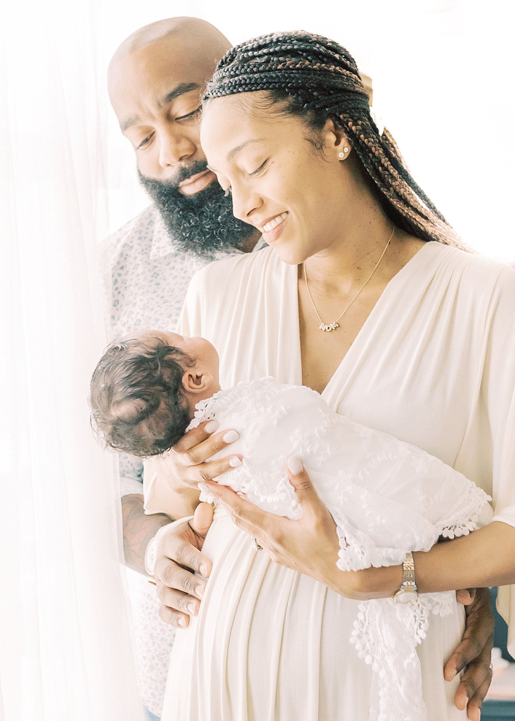 man and woman holding a newborn baby girl in front of a light and airy window wrapped in white