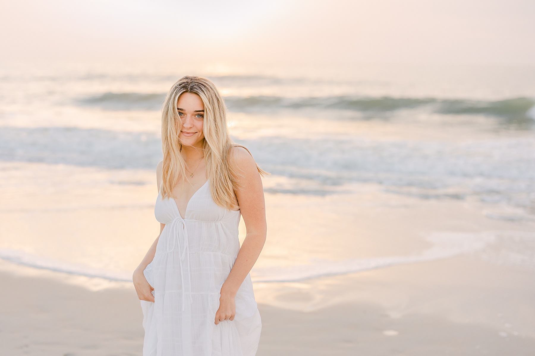 blond woman standing on the beach at sunrise in white maxi dress