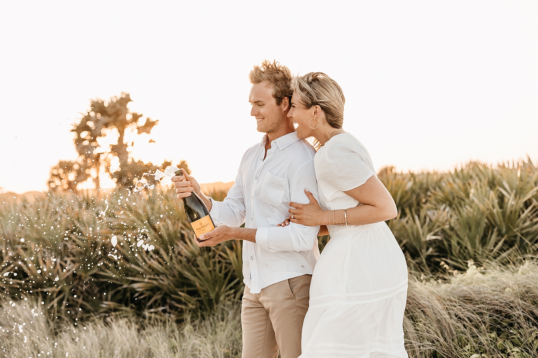 blond hair woman long white dress on the beach popping bottle of champagne with man in khaki pants