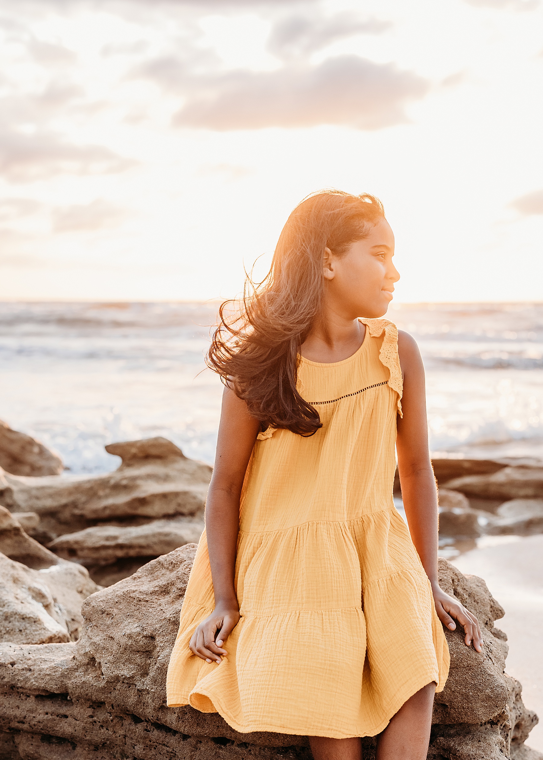 little girl sitting on the rocks on the beach at sunrise in yellow dress