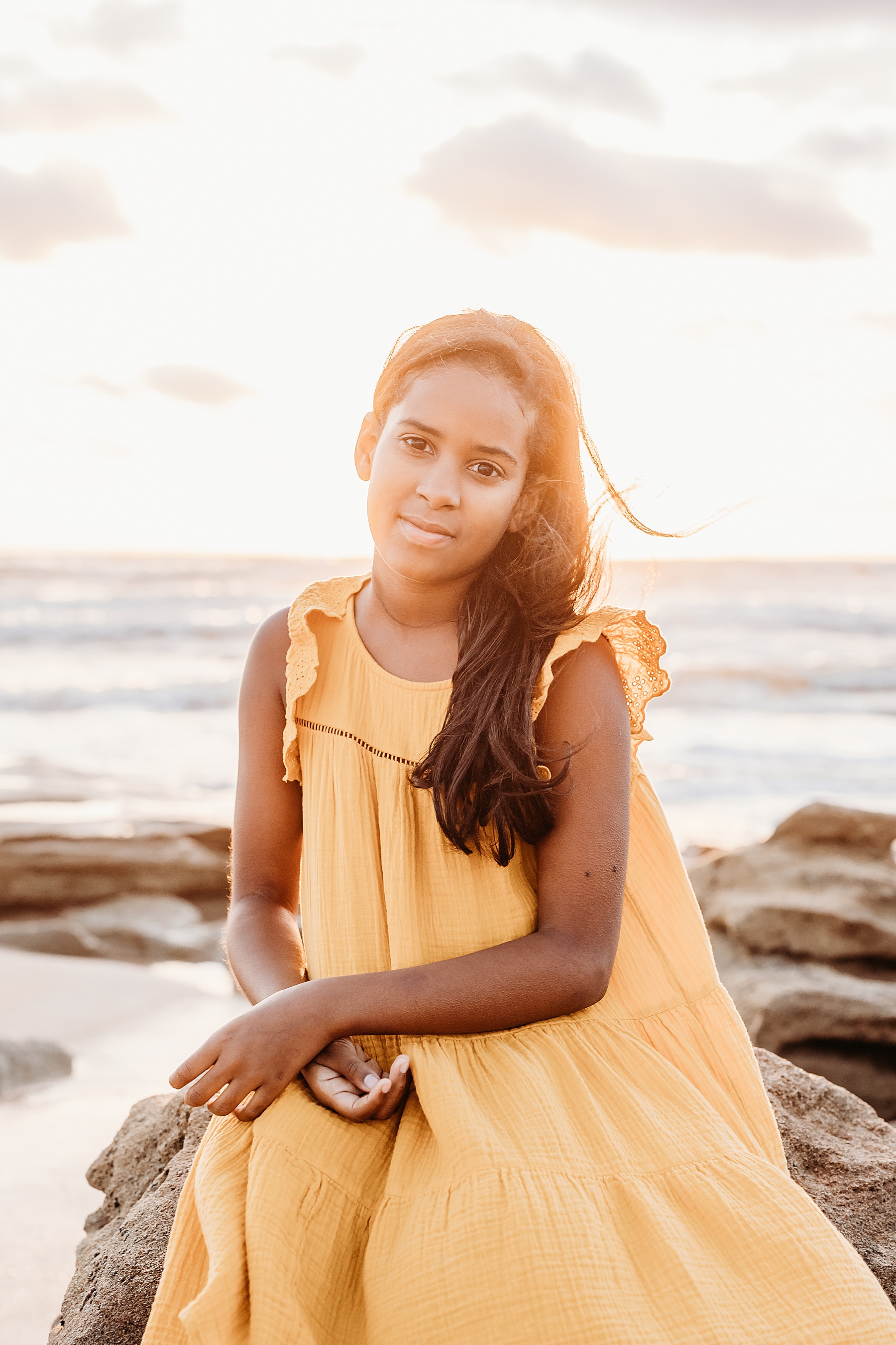 little girl wearing yellow dress sitting on the rocks on the beach at sunrise