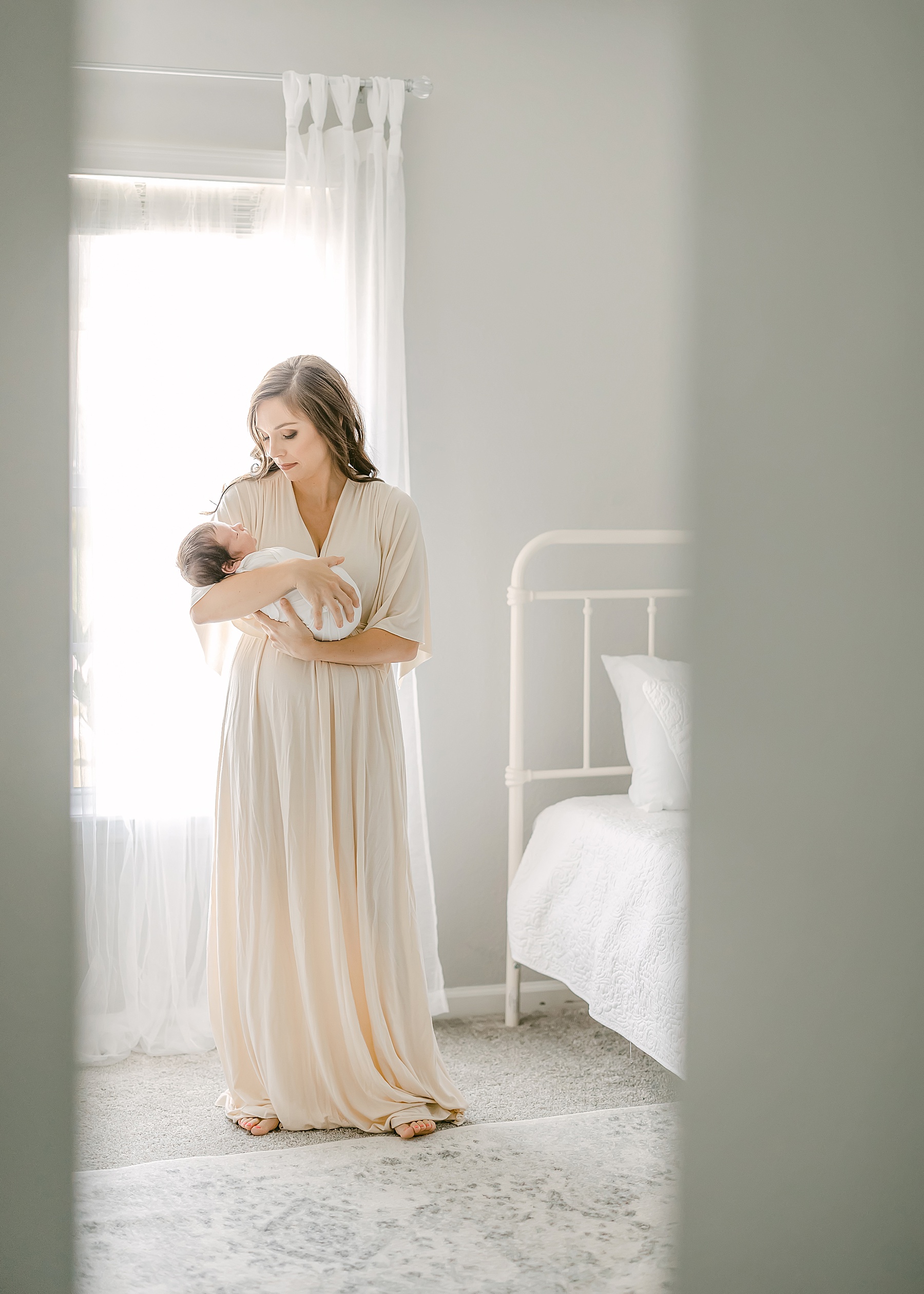 woman in cream maxi dress holding newborn baby boy against gray wall in neutral nursery 