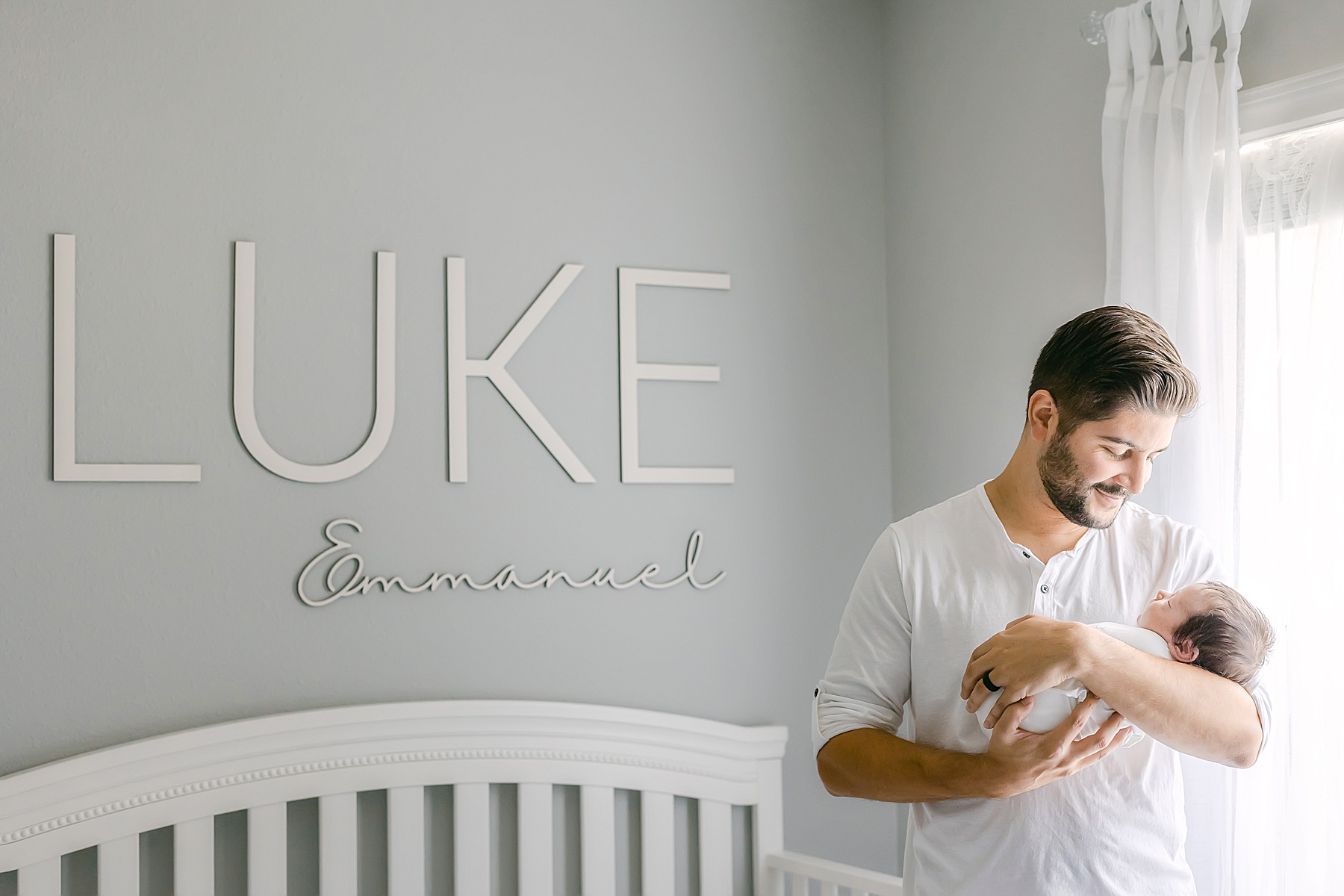 man in white shirt holding newborn baby boy against gray wall in nursery