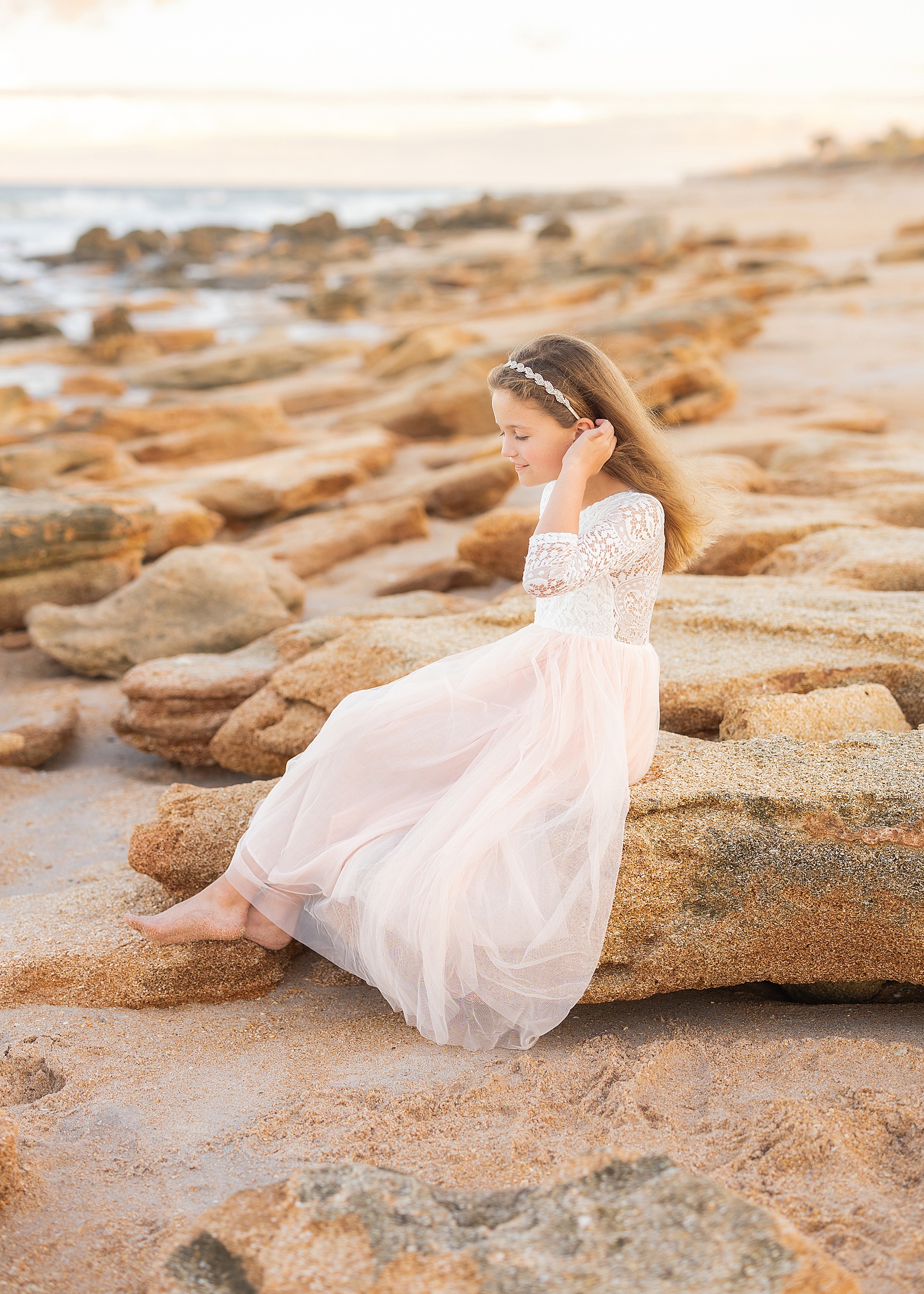 little girl wearing white and tulle dress sitting on the rocks at sunset in saint augustine beach florida