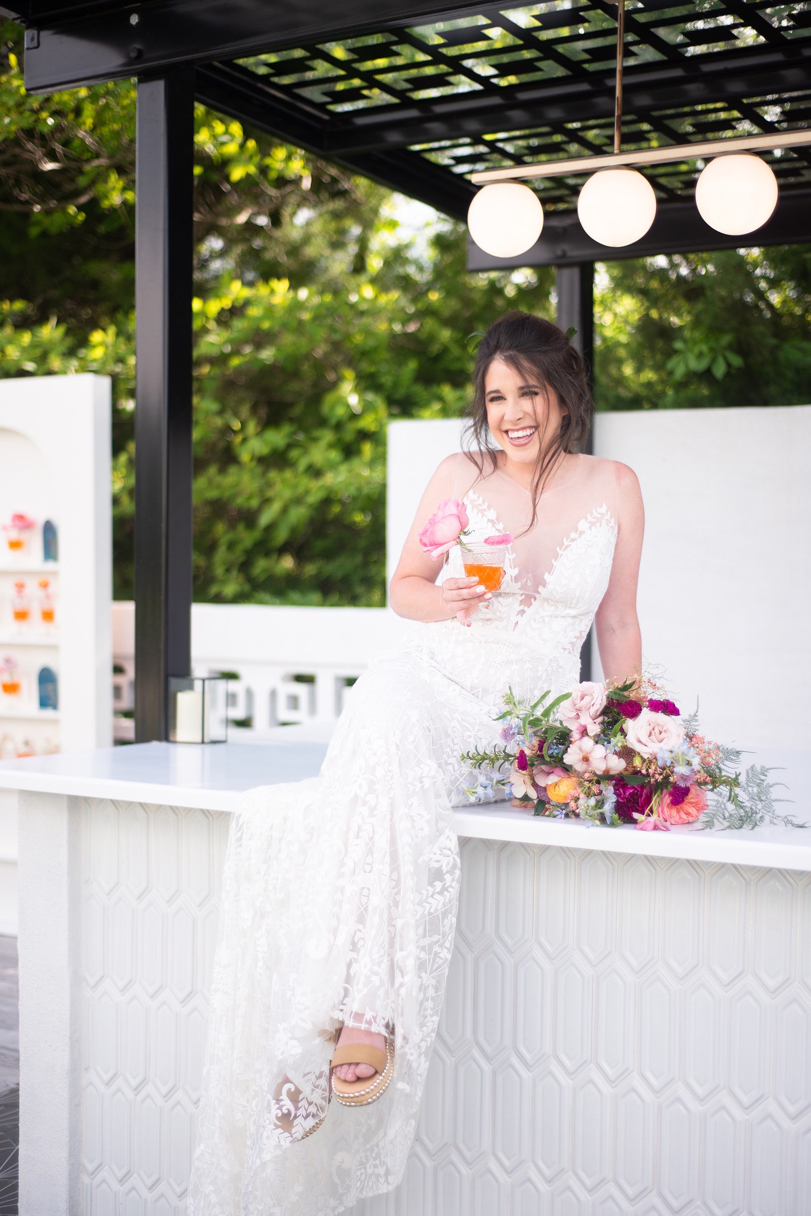Bride sitting on outdoor bar at Greenhouse Two Rivers with an orange drink.