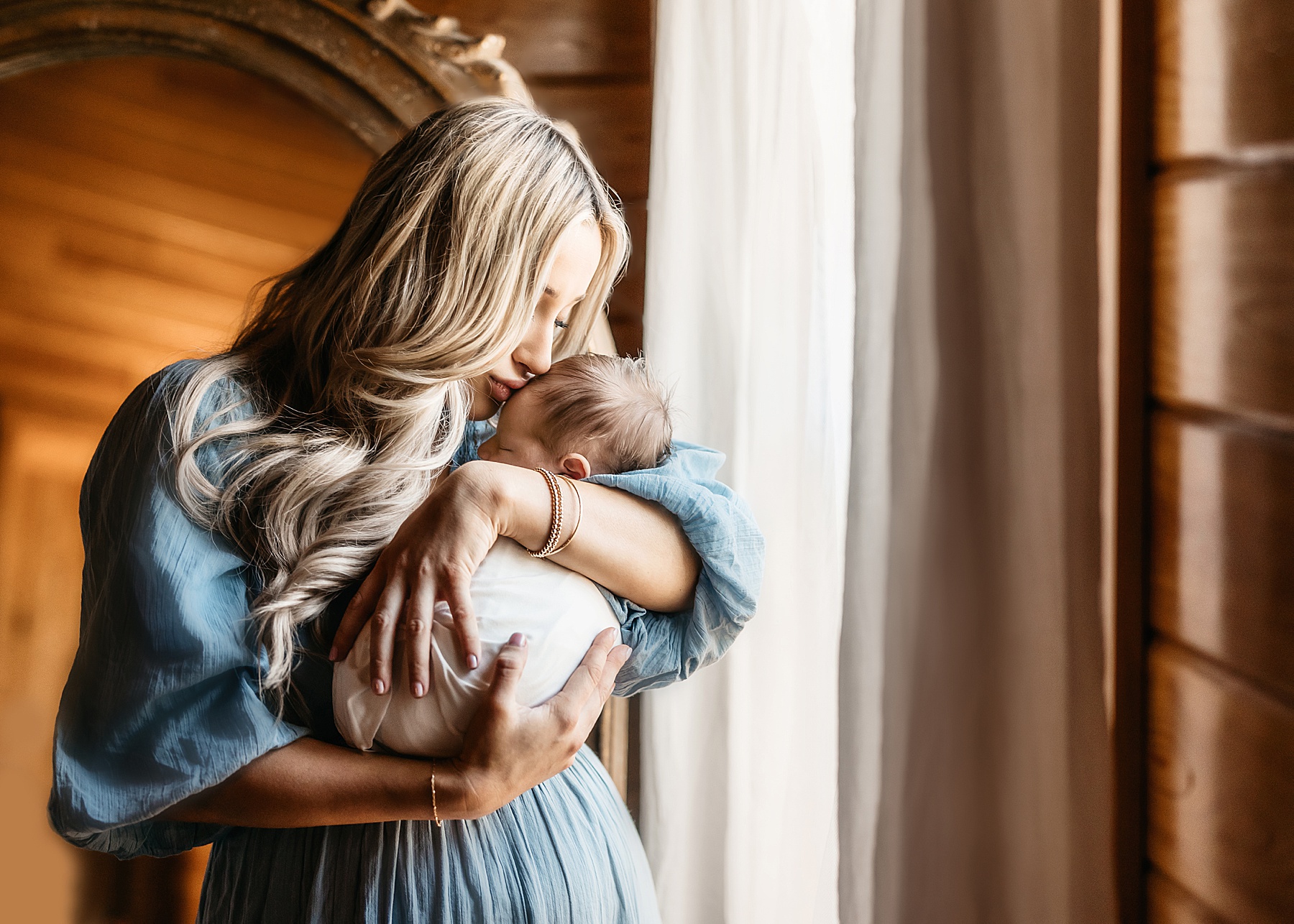 woman with long blond hair kissing baby boy wrapped in white swaddle