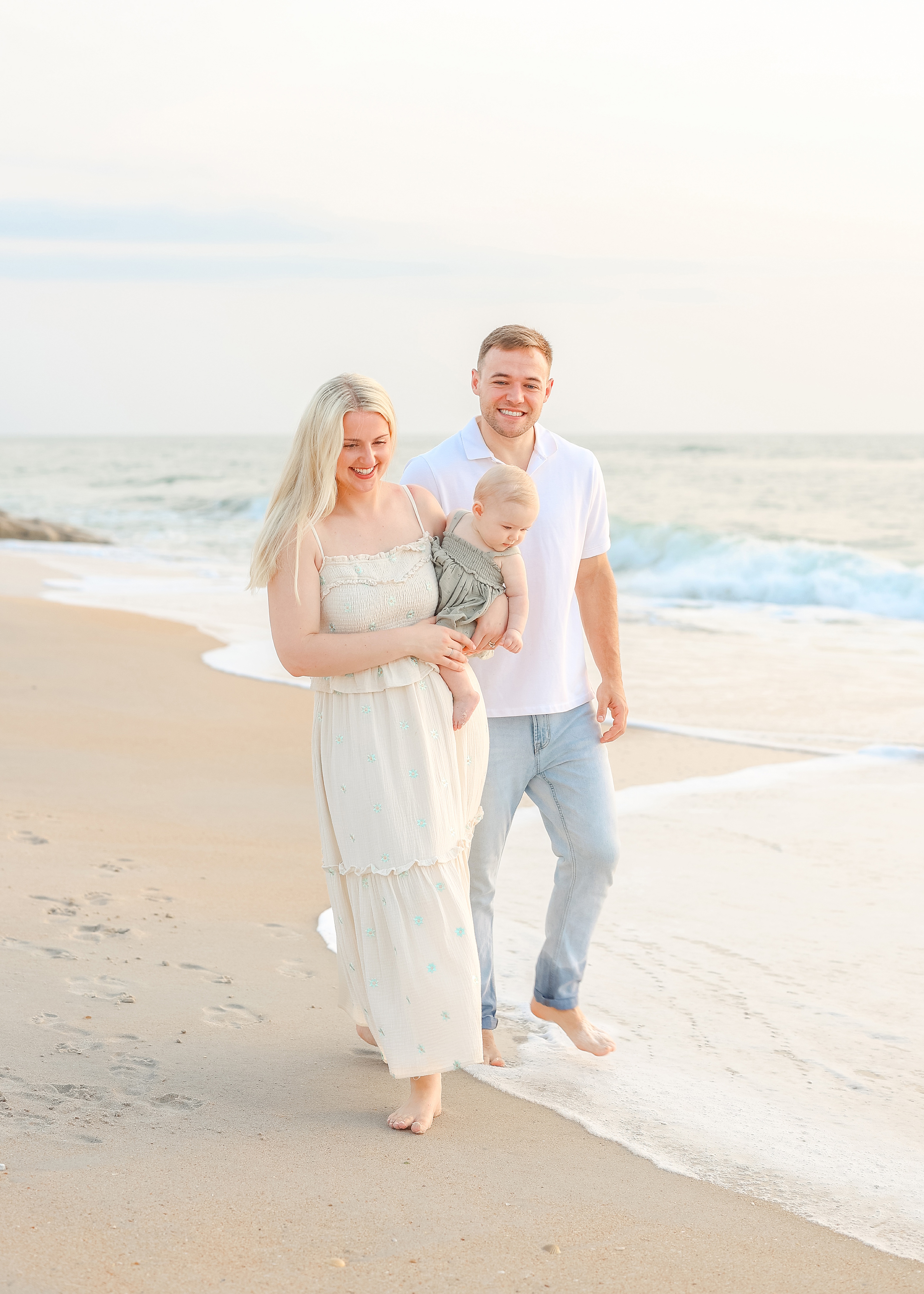 family walking near the shoreline at the beach holding baby girl in white dress
