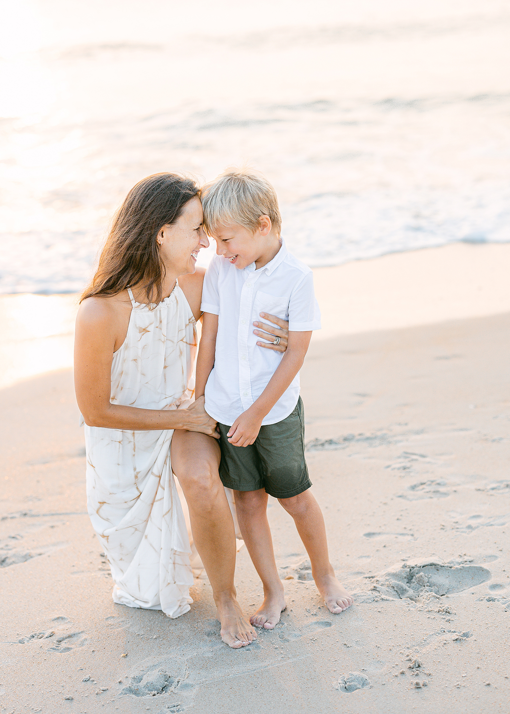 Woman in white dress holding little boy on the beach at sunrise.