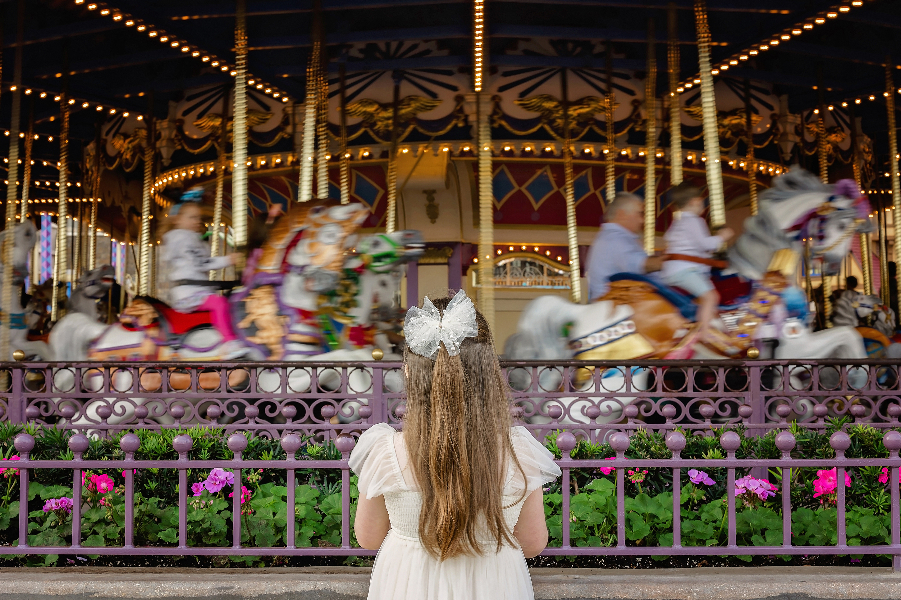 A little girl with a cream bow in her hair looks onto a colorful moving carousel. 