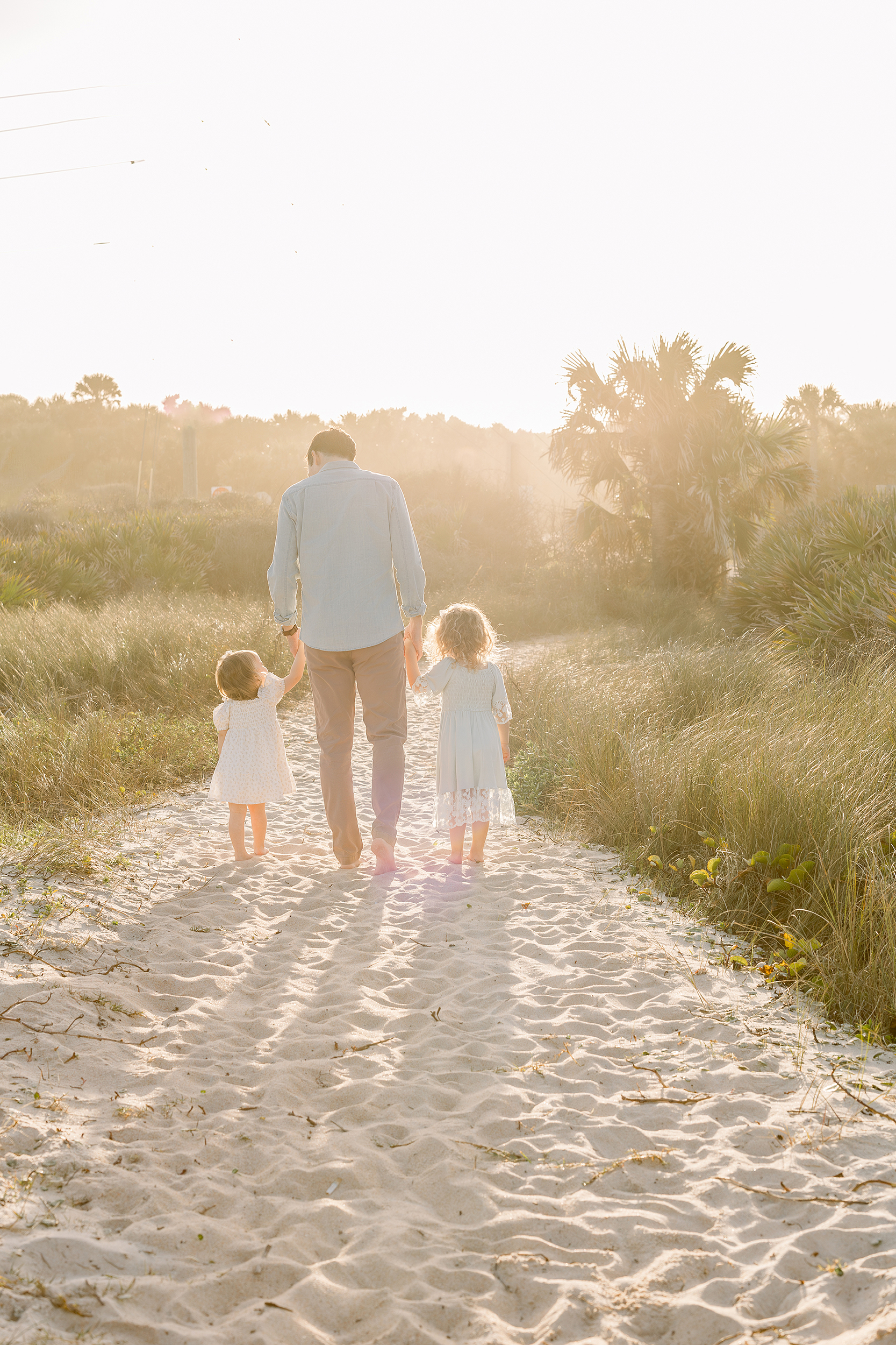 A light filled beach portrait of a father walking down a sand path with his two little girls at sunset.