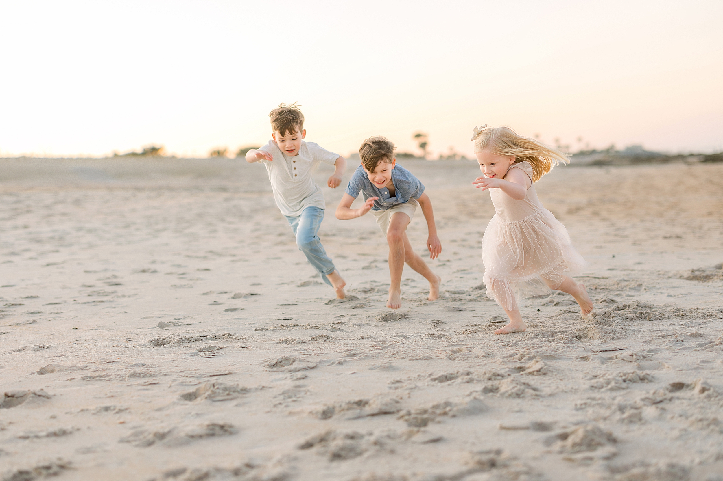 Three children run on the beach together at sunset.