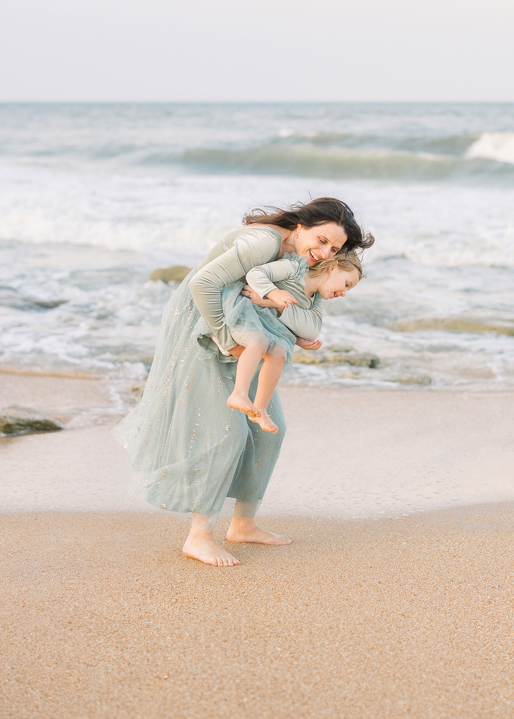 A mother swings her daughter by the water at the beach at sunset.