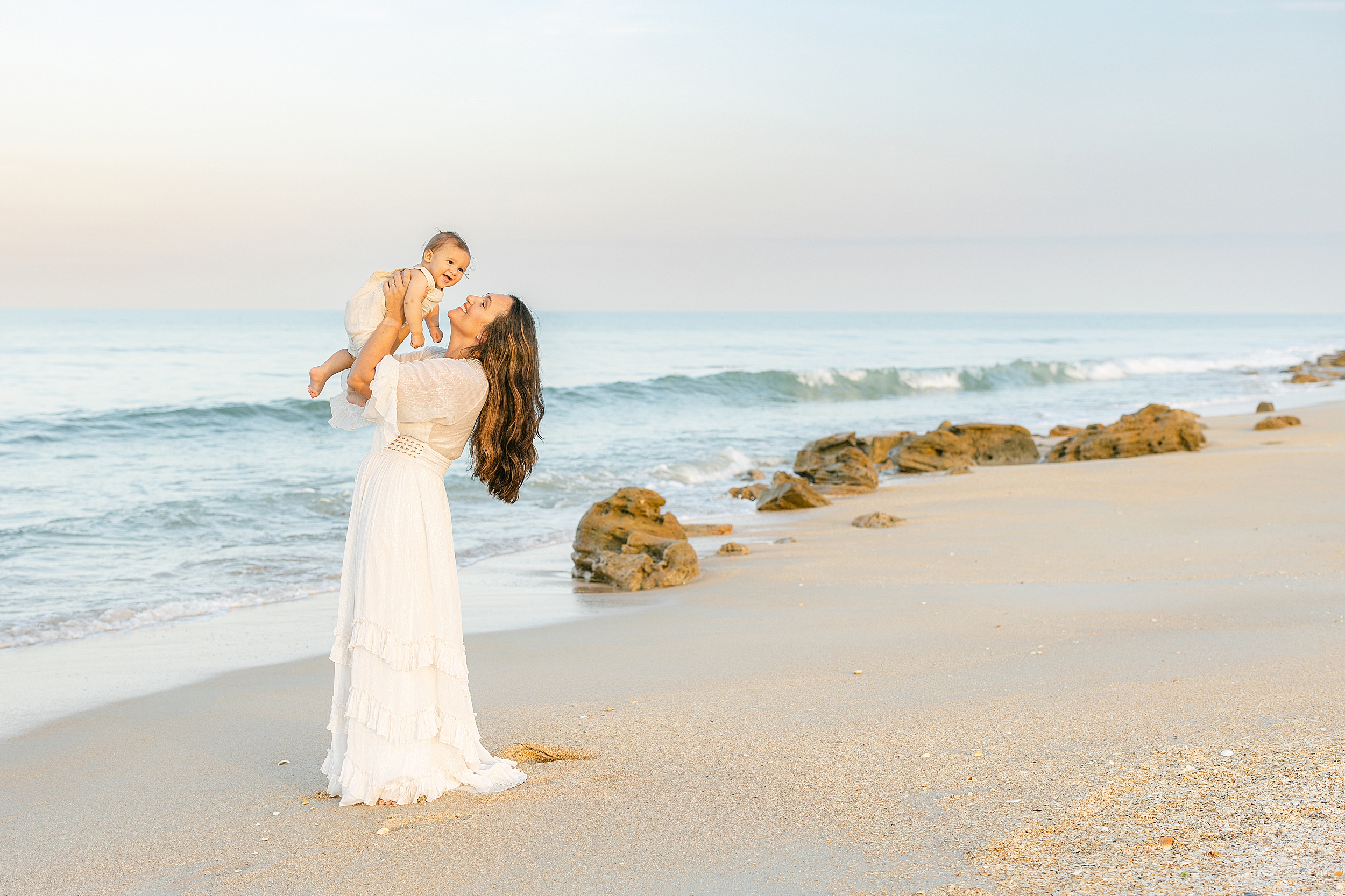 A woman holds her baby boy up in the air on the beach during a colorful sunset.