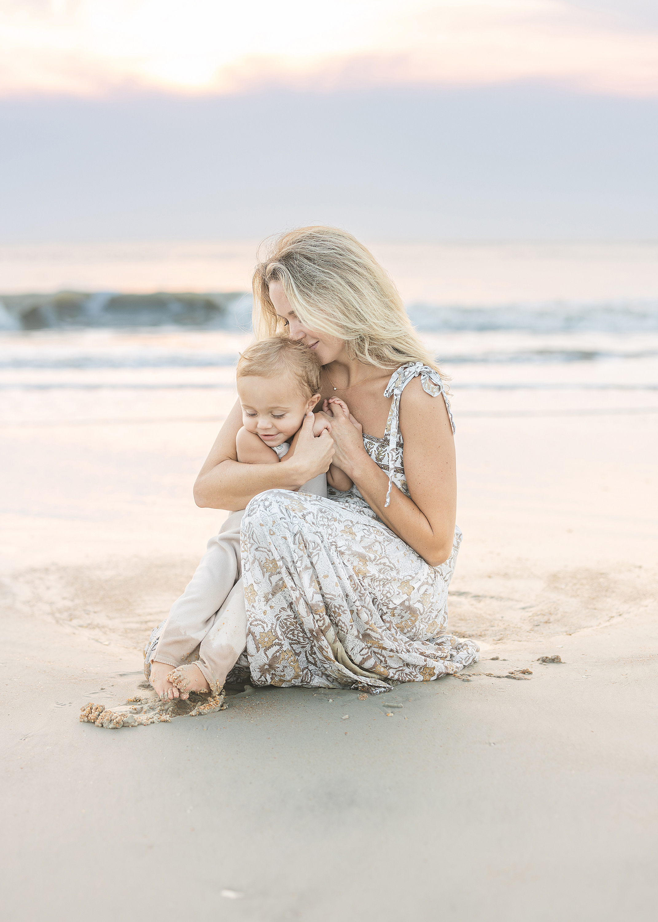 A pastel colored sunrise beach portrait of a woman sitting on the sand at the beach with her baby boy.