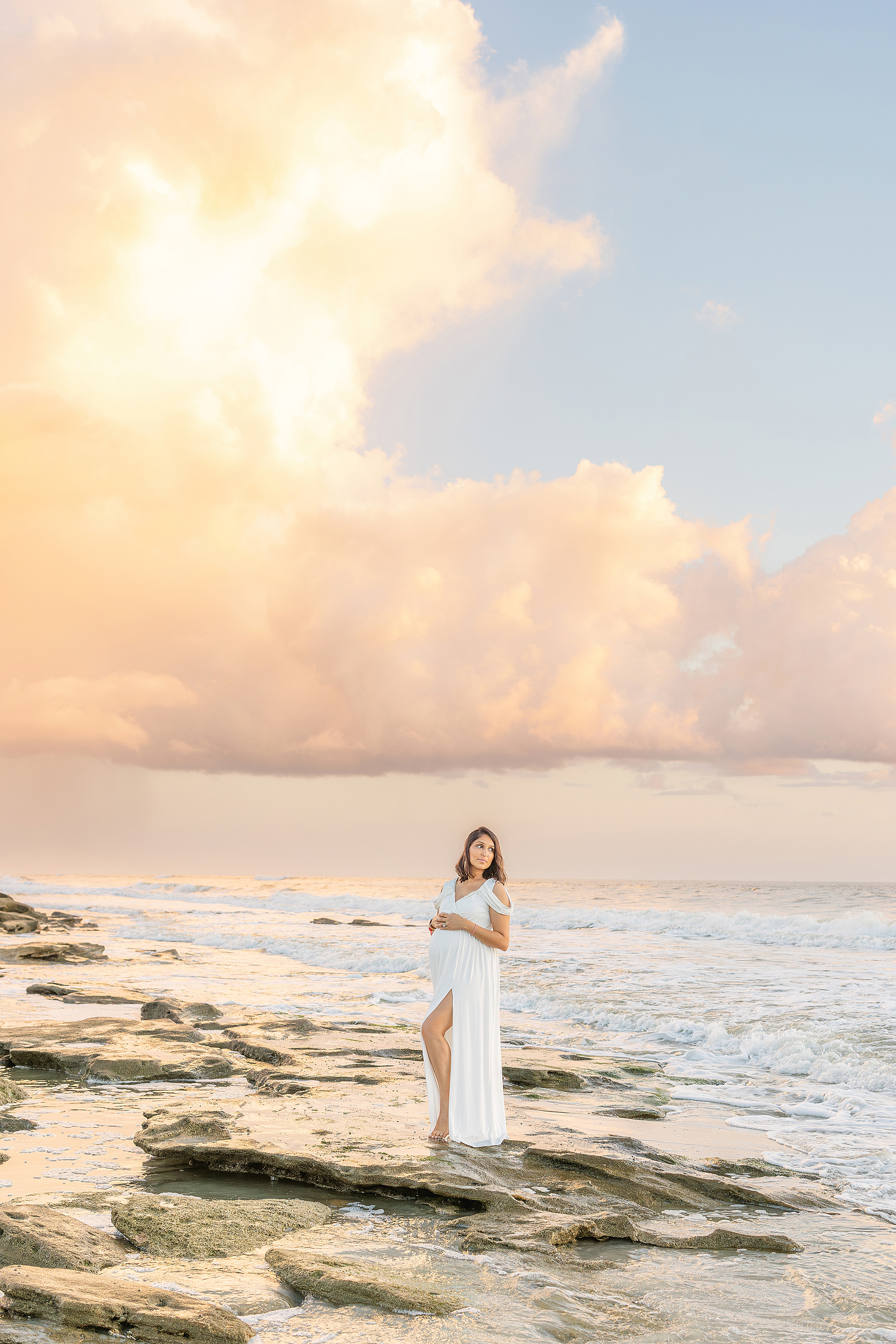 A sunrise maternity portrait of a woman in a long white dress on the rocks at Saint Augustine Beach at sunrise.