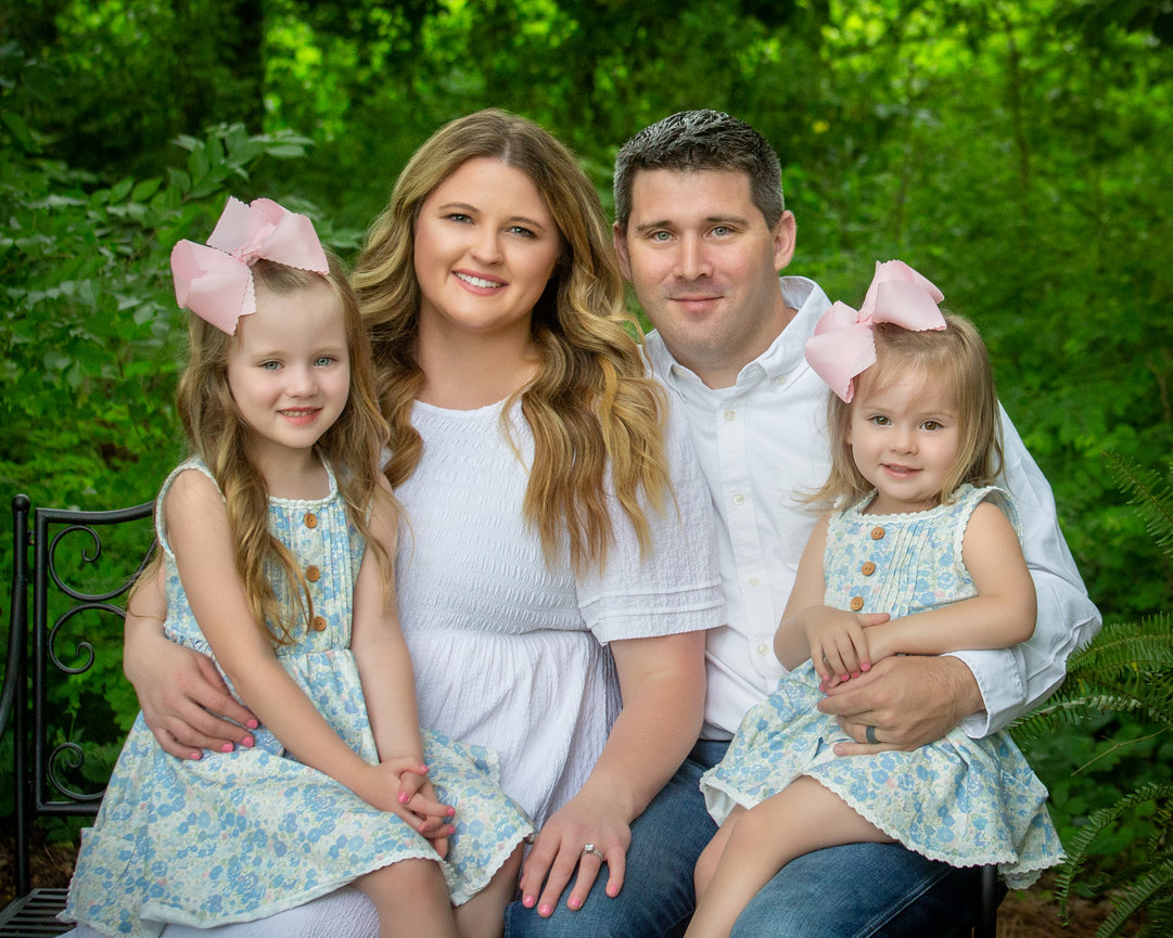 A family sitting together on a bench in a garden, with parents and two young girls wearing matching dresses and pink bows.