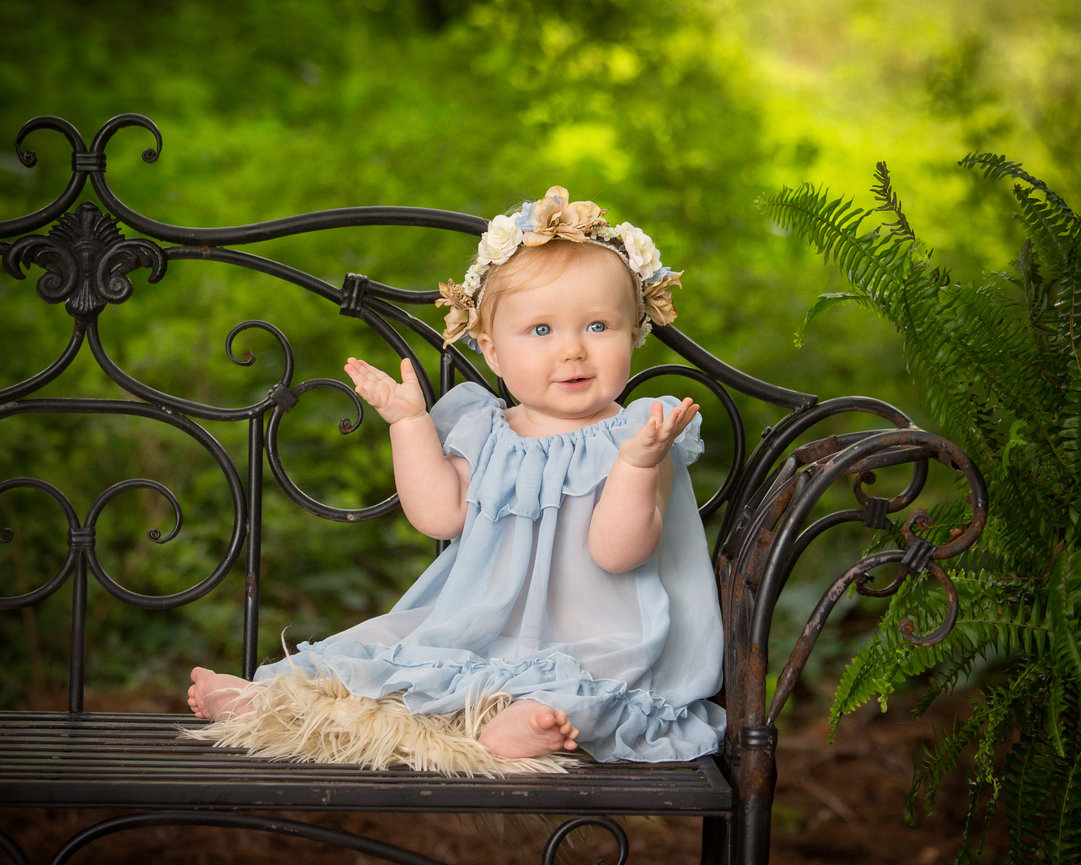 Baby in blue dress and floral headband sitting on a wrought iron bench outdoors, surrounded by greenery.