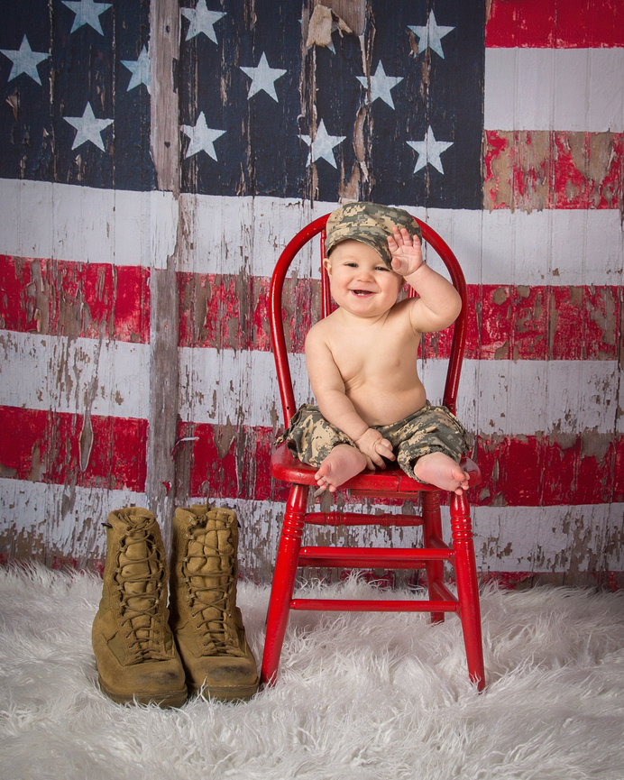 Baby in camouflage sits on a red chair, waving, with American flag backdrop and military boots nearby.