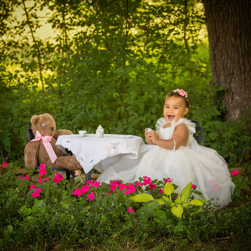 Toddler in a white dress having a tea party with a teddy bear in a garden setting.