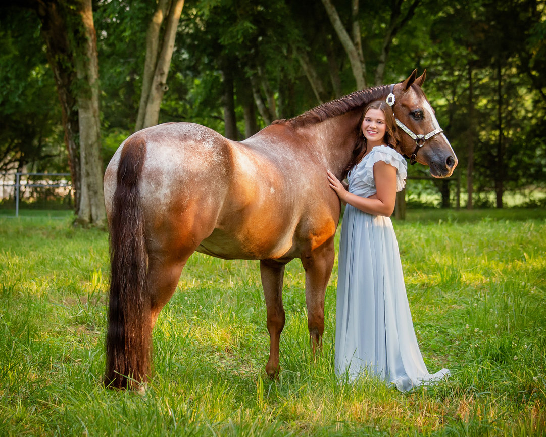 Woman in a light blue dress stands next to a brown horse in a grassy area with trees.