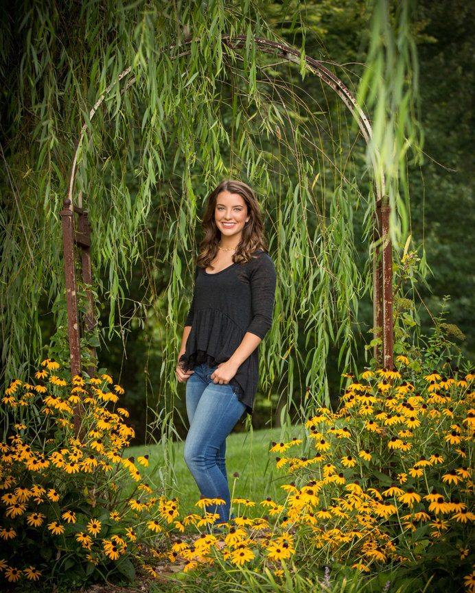 Woman in a black top and jeans standing under a willow archway surrounded by yellow flowers.