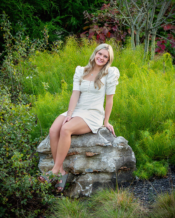 Woman in a white dress sitting on a rock surrounded by greenery and vibrant plants.