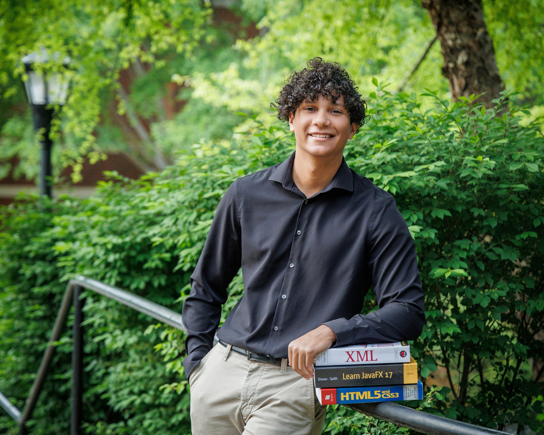 Young man in a black shirt leaning on a railing, smiling while holding programming books in a green outdoor setting.