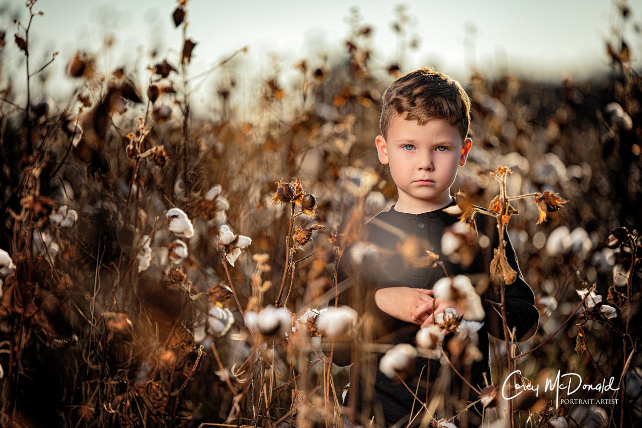 Child standing amidst a field of dried cotton plants, wearing a dark shirt, with a serious expression.