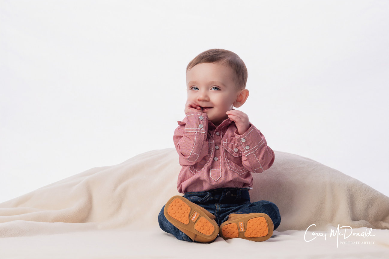 A toddler in a pink shirt and jeans sits on a soft blanket, smiling with hands near face.