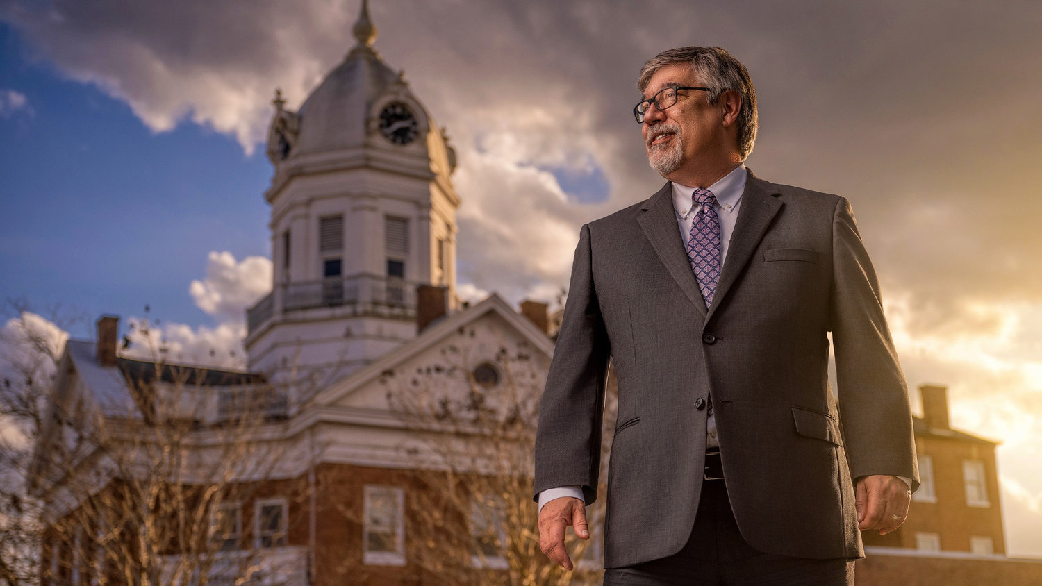 political man wearing a suit and tie standing outside monroeville courthouse at sunset with dramatic lighting and skies from ground perspective