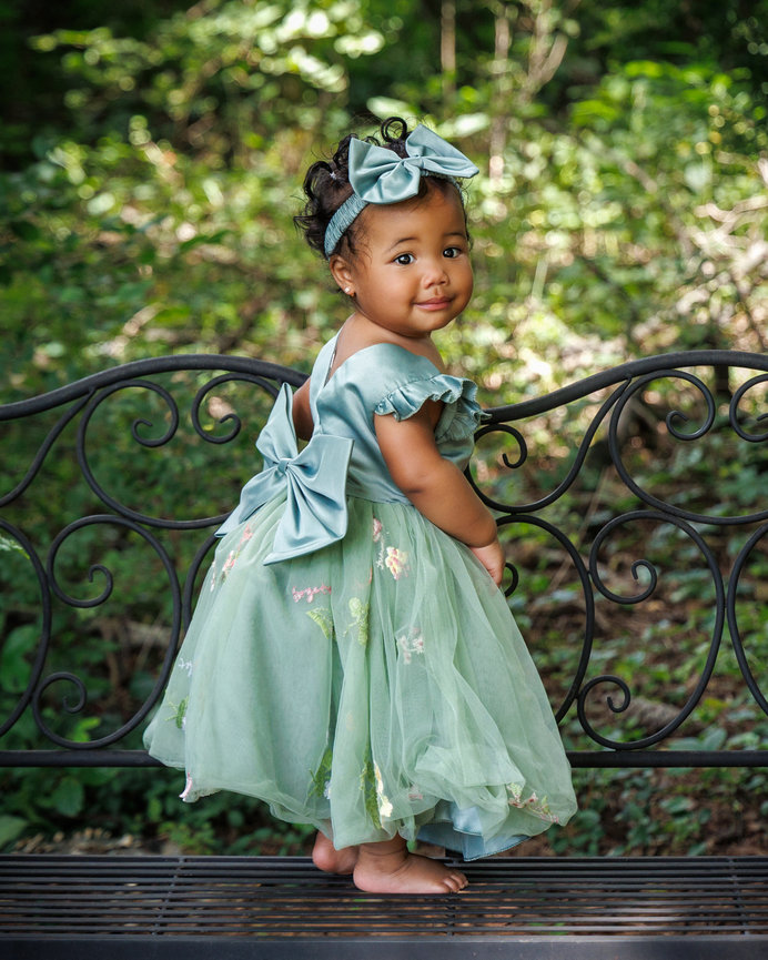 A toddler in a green dress and bow stands on a decorative bench in a lush, leafy garden.