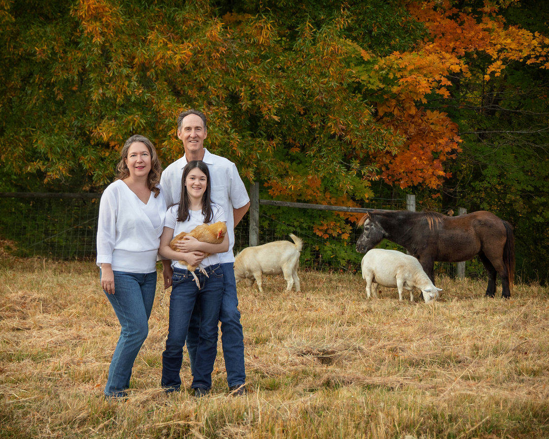 Family in a field with goats, a horse, and autumn trees in the background. One person holds a chicken.