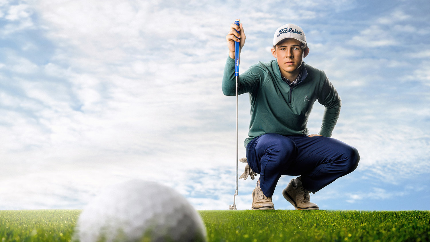 Golfer crouching on green, focusing on a golf ball, with blue sky and clouds in the background.
