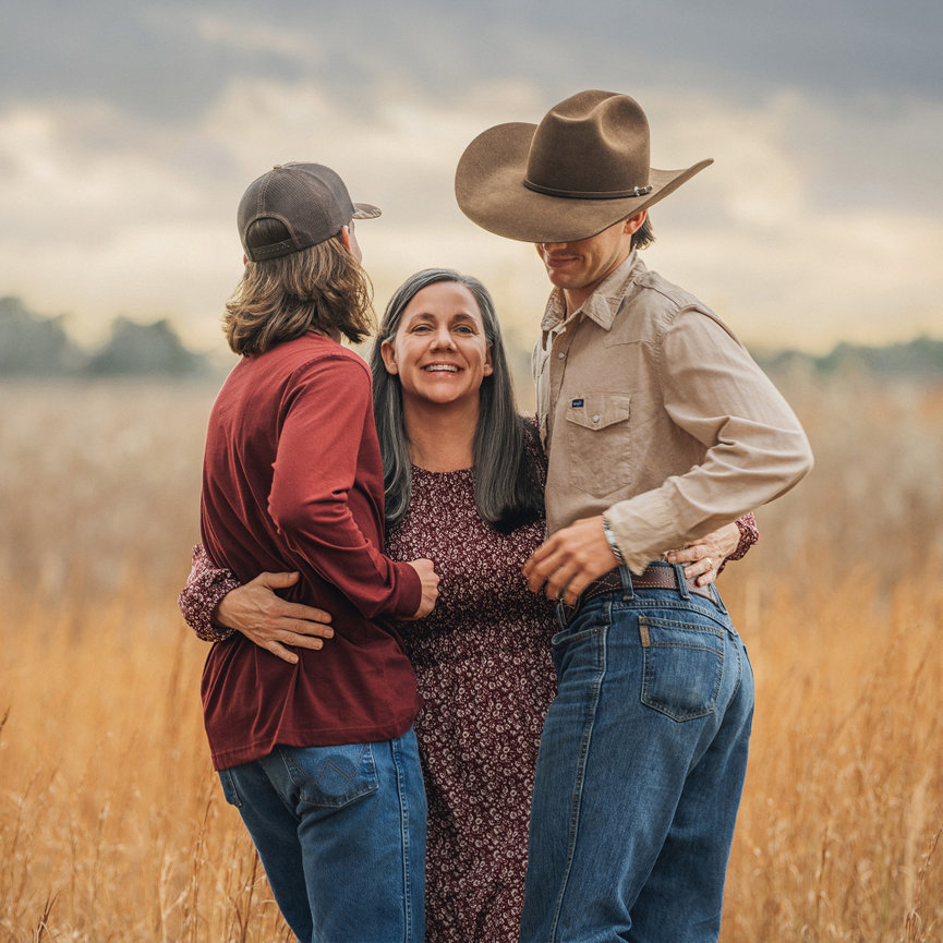 A smiling woman stands between two men in a field, all embracing. The men wear casual clothing and cowboy hats.