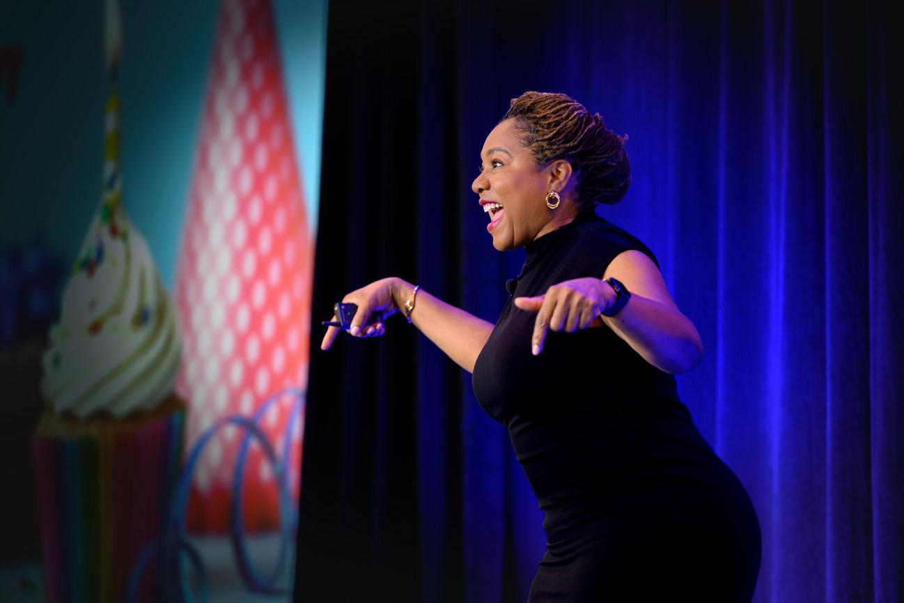 Smiling woman speaking on stage with blue lighting and gesturing with both hands