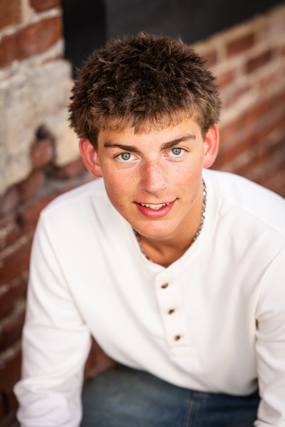Franklin area senior picture of a teenage guy smiling at the camera with a white long sleeve shirt and jeans on in front of a brick background by Seniority Nashville