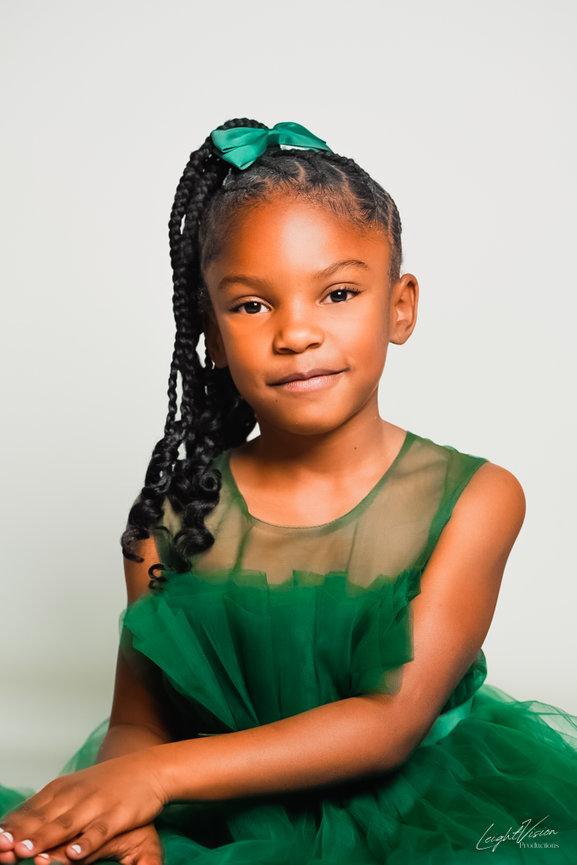 A young girl wearing a green dress stands confidently, posing for a photo with a bright smile on her face.