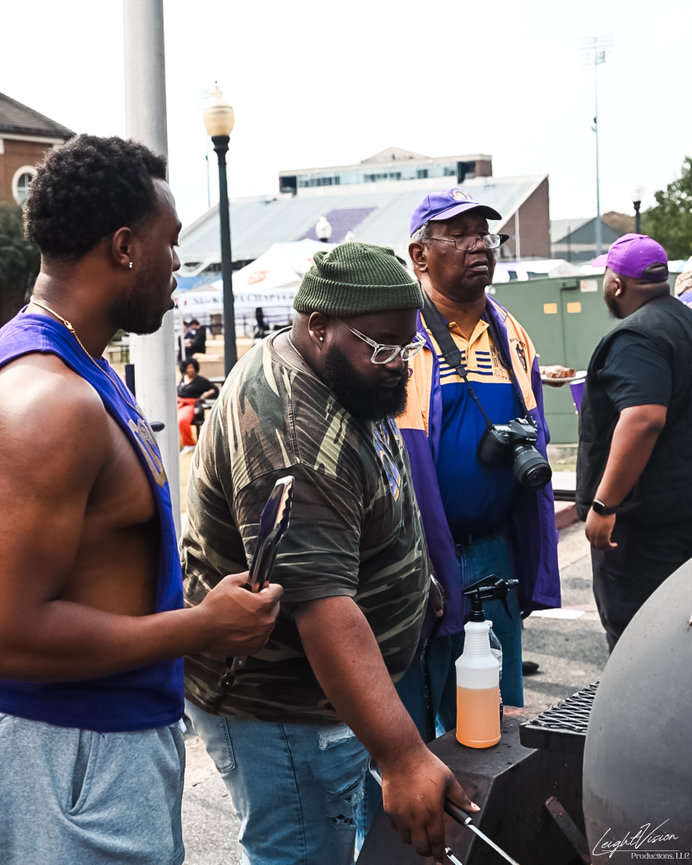 A gathering of men around a grill, actively cooking and socializing in an outdoor environment.