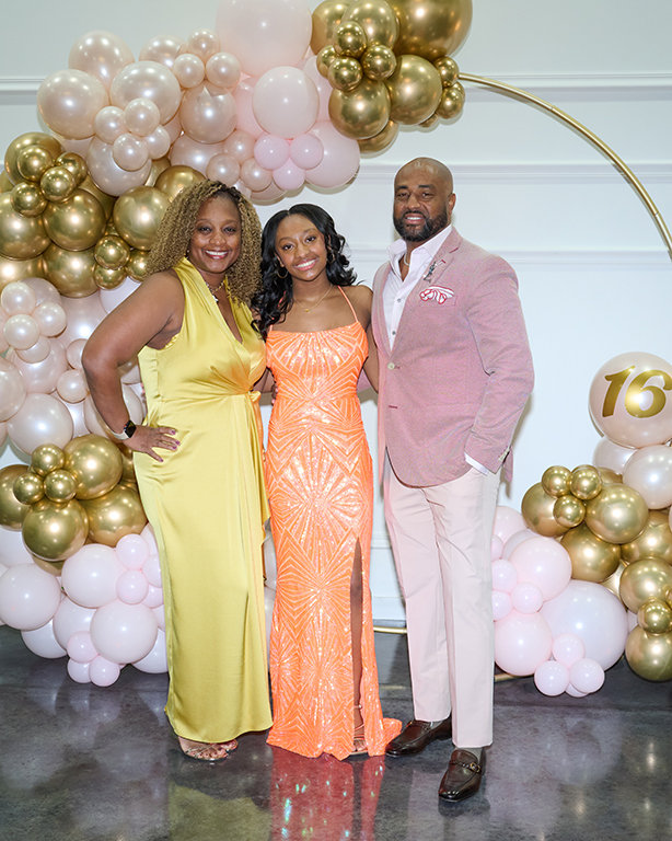 Three people posing in colorful formal attire at a balloon-decorated event backdrop.