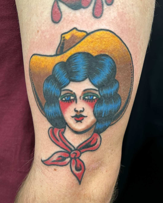 Blue haired cowgirl tattoo by Beam. The blue eyed cowgirl is wearing a yellow cowboy hat and has a red scarf tied around her neck. The tattoo is done in the traditional tattooing style giving it an old school vintage feel.