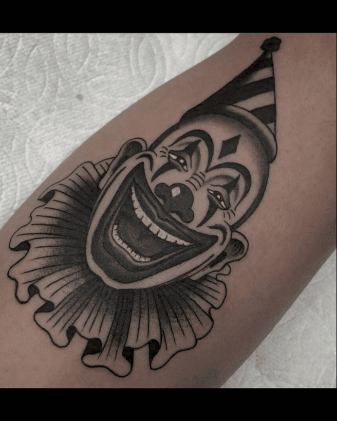 Black and grey tattoo of a smiling clown with striped hat and frilled collar.