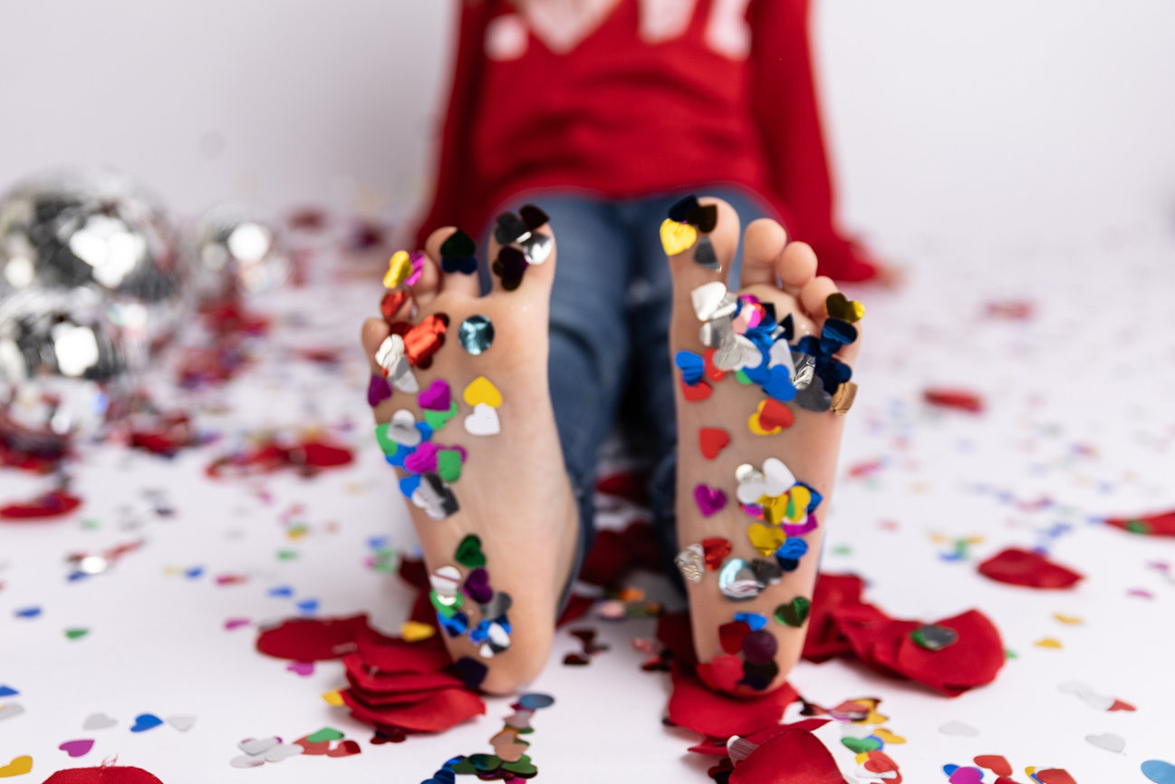 Child seated with feet covered in colorful heart-shaped confetti, surrounded by scattered confetti and red rose petals.
