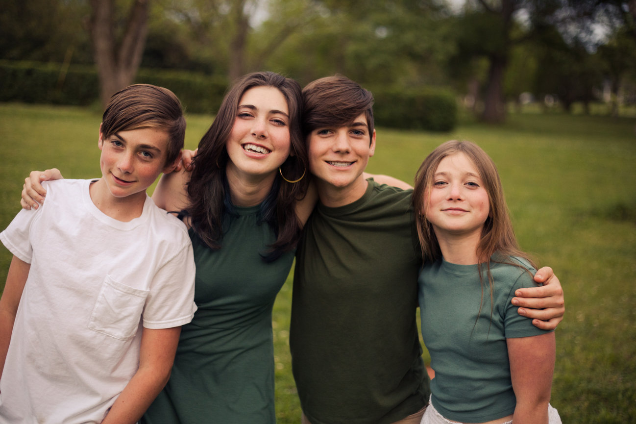 Four siblings standing shoulder to shoulder and smiling outdoors.