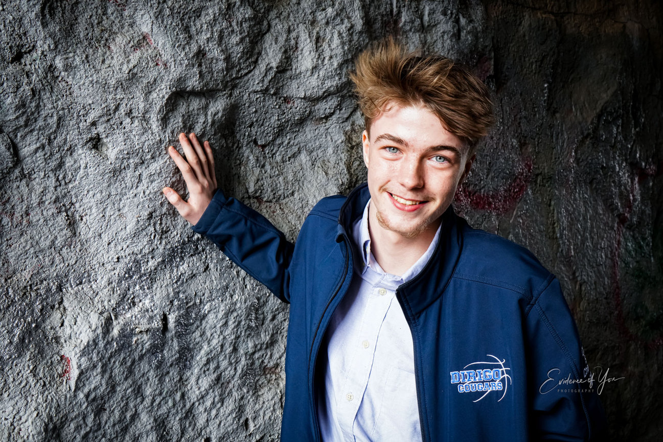Young person smiling in a blue jacket, standing against a textured stone wall.
