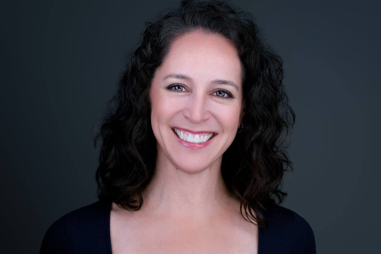 Smiling woman with dark curly hair in studio headshot on gray background, taken by professional DFW portrait photographer