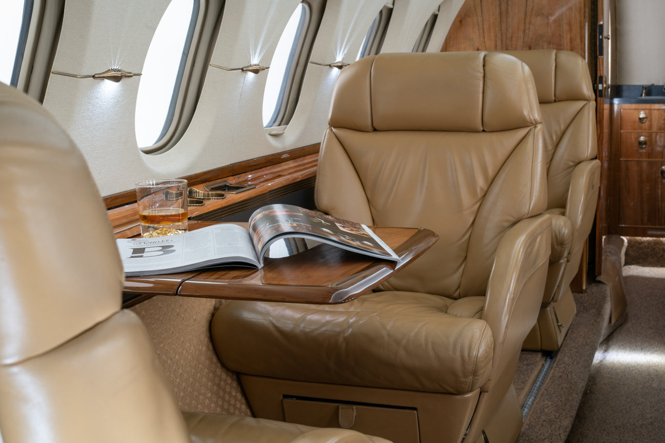 Luxury private jet interior with leather seats, a wooden table, an open magazine, and a glass of whiskey.