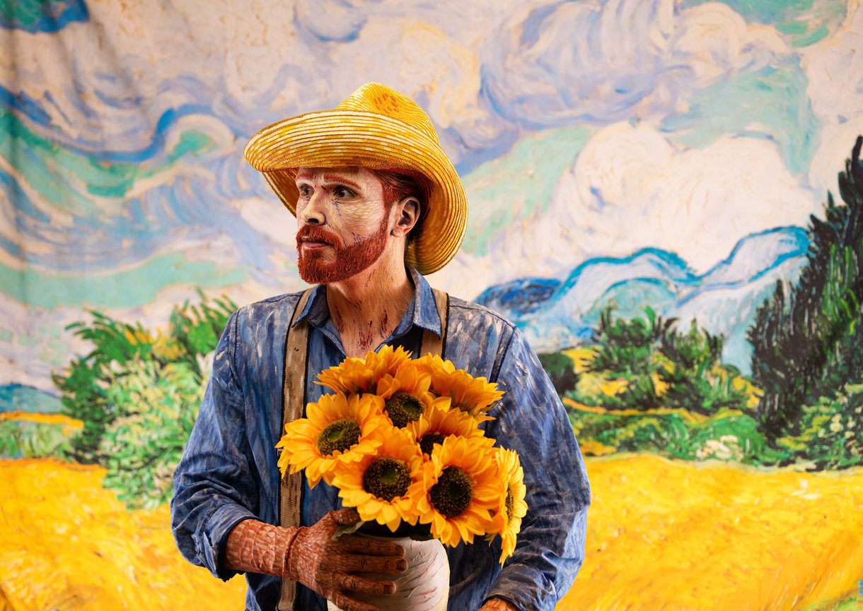 Man with a hat holding sunflowers standing in front of a painted landscape