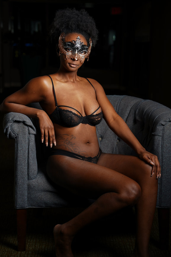 A woman in a black lace lingerie set and mask sits elegantly on a gray chair in dim lighting.